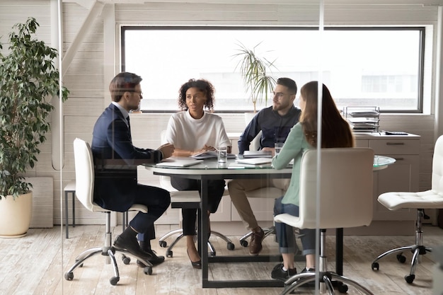 Diversos socios de negocios discutiendo los términos del contrato del proyecto haciendo una lluvia de ideas en la sala de juntas moderna detrás de los colegas de la pared de vidrio sentados en la mesa en la oficina hablando compartiendo ideas de inicio