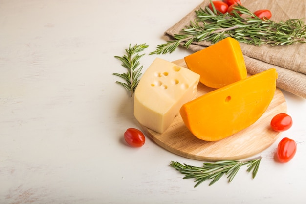 Diversos tipos de queso con romero y tomates sobre tabla de madera sobre una superficie de madera blanca y textiles de lino