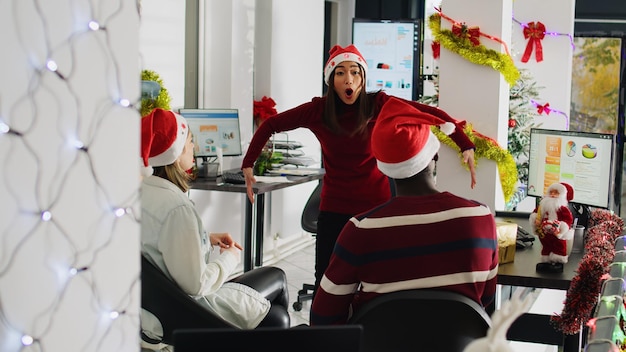 Diversos trabajadores con sombrero de Papá Noel disfrutando del espíritu navideño jugando a simular un juego de actuación durante las vacaciones. Compañeros de trabajo divirtiéndose haciendo gesticulaciones divertidas durante la pantomima en Navidad adornan la oficina