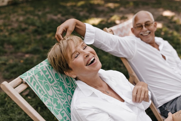 Divertida anciana con peinado corto en blusa blanca riendo y divirtiéndose con un marido sonriente en camisa ligera al aire libre