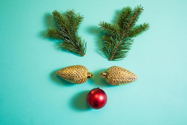 Divertida cara de ciervo hecha de artículos relacionados con la Navidad juguetes dorados del árbol de Navidad en forma de conos de pino ramas de abeto y bola roja Concepto minimalista Sobrecarga sobre fondo verde claro