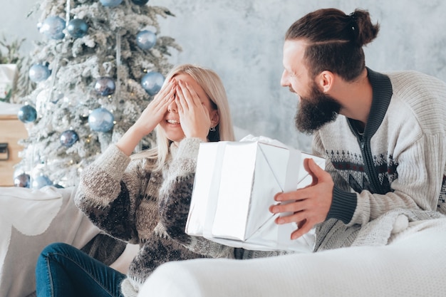 Divertida celebración navideña. Guy preparó un regalo sorpresa para su novia. Señora emocionada por el presente, cubriéndose los ojos, sonriendo.