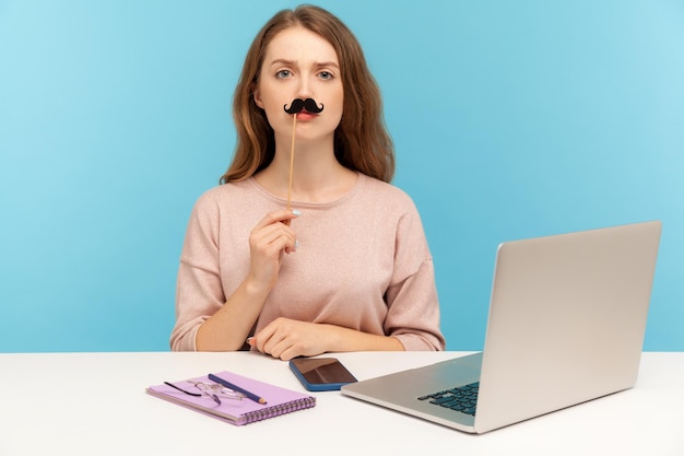 Divertida empleada de oficina seria sentada en el lugar de trabajo cubriendo los labios con bigote de papel falso imitando al jefe estricto accesorio de mascarada humorística tiro de estudio interior aislado en fondo azul