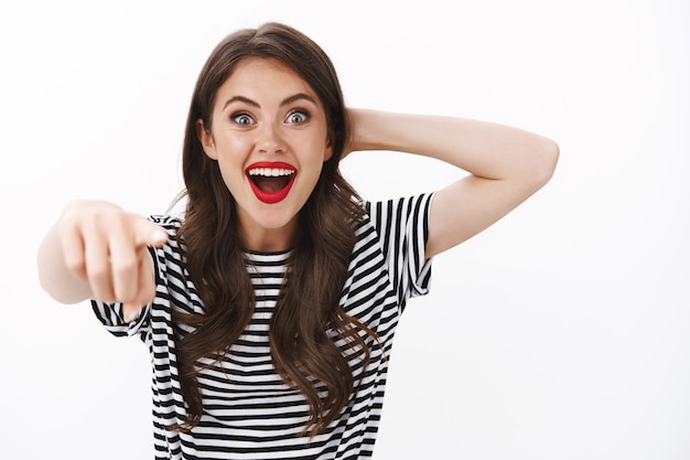 Divertida y excitada mujer seductora linda con corte de pelo rizado y tatuaje, agarrar la cabeza asombrada, apuntando a la cámara, mirada emocionada, emboscada, feliz de ver al actor favorito, de pie sobre una pared blanca emocionada