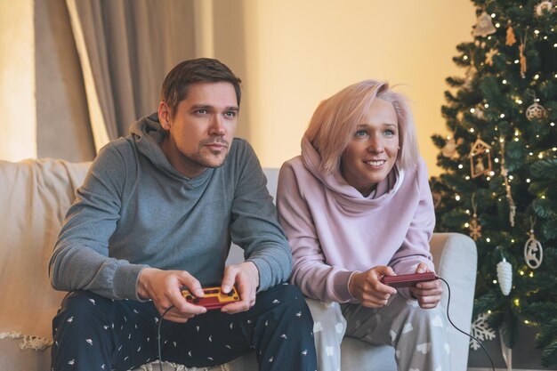 Divertida familia joven vestida de casa jugando videojuegos en el sofá al fondo del árbol de Navidad