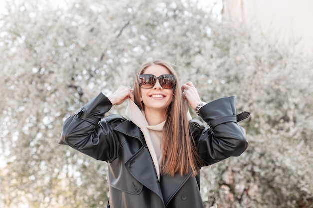 Divertida, feliz y hermosa modelo de chica de moda con una sonrisa con gafas de sol en un abrigo negro de moda con una capucha, usa una capucha, disfruta y camina en un parque floreciente