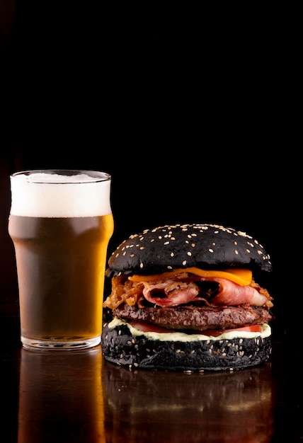 Divertida hamburguesa con temática de halloween panecillo negro crujiente tocino y rodajas de cebolla con sésamo en una mesa de madera servida con media pinta de cerveza artesanal retrato