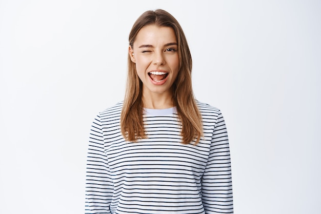 Divertida joven rubia guiñando un ojo y sonriendo, expresa alegría y emociones positivas, de pie sobre una pared blanca en una camisa a rayas casual