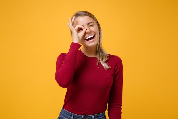 Divertida jovencita rubia con ropa informal posando aislada en un fondo amarillo anaranjado de la pared. Concepto de estilo de vida de las personas. Simulacros de espacio de copia. Sosteniendo la mano cerca del ojo, imitando binoculares o anteojos.