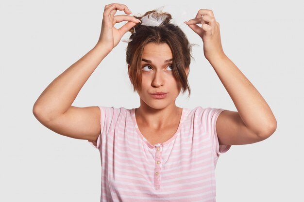 Divertida juguetona jovencita juega con plumas en la cabeza, usa ropa casual, posa contra la pared blanca en el dormitorio, no tiene nada que hacer después de un largo sueño. Concepto de personas, hora de dormir, descanso y fin de semana