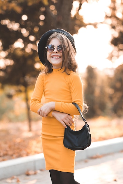 Divertida y linda niña de 4 a 5 años que usa gafas de sol y un vestido de punto amarillo posando sobre la naturaleza en otoño