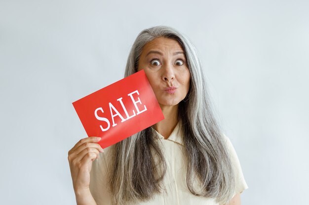 Foto divertida mujer asiática de mediana edad con cabello plateado suelto muestra cartel de venta rojo haciendo muecas sobre fondo claro en estudio