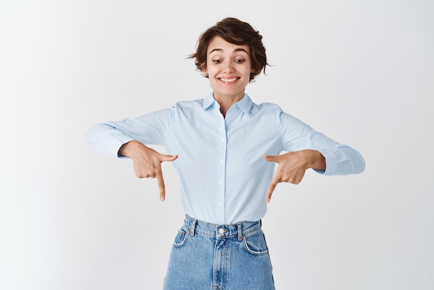 Divertida mujer feliz con camisa de cuello azul mirando y señalando con el dedo hacia abajo con una sonrisa asombrada y alegre de pie sobre fondo blanco