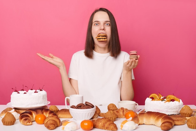 Divertida mujer hambrienta de cabello castaño con camiseta blanca sentada en la mesa con dulces aislados sobre fondo rosa posando con la boca llena de galletas