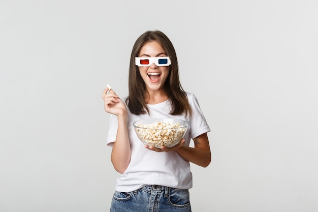 Divertida mujer joven y bonita con gafas 3d viendo películas o series de televisión, comiendo palomitas de maíz y sonriendo emocionado.