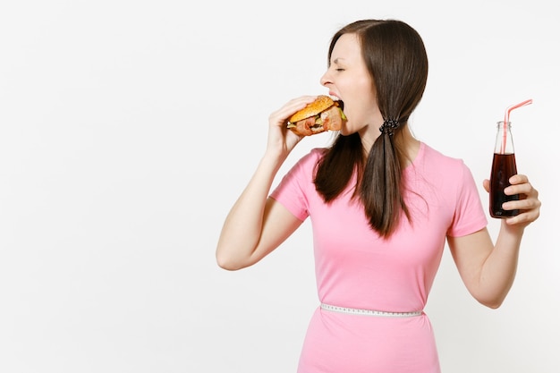 Divertida mujer joven con colas, cinta métrica en la cintura sosteniendo hamburguesa, cola en botella de vidrio aislada sobre fondo blanco. Nutrición adecuada o comida rápida clásica estadounidense. Área de publicidad con espacio de copia.