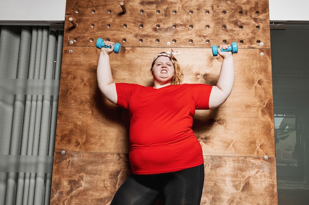 Divertida mujer con sobrepeso vestida con ropa deportiva y con un vendaje en la cabeza trabaja con pesas en el fondo de una pared de madera en el gimnasio