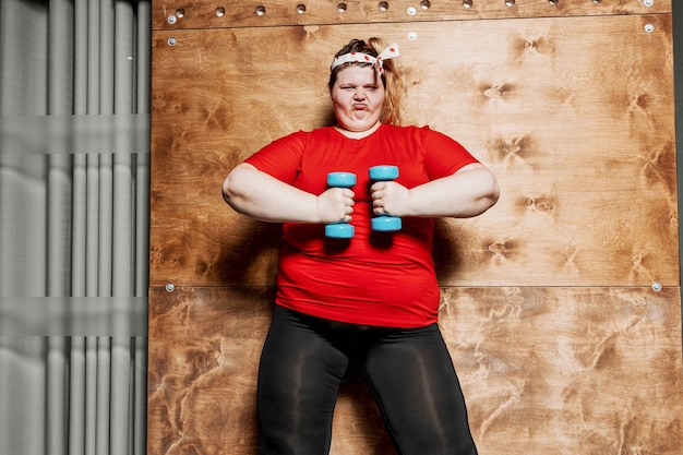 Divertida mujer con sobrepeso vestida con ropa deportiva y con un vendaje en la cabeza trabaja con pesas en el fondo de una pared de madera en el gimnasio