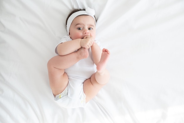 Divertida niña de 6 meses jugando con las piernas, acostado en la cama en su casa en ropa de cama blanca. vista superior.