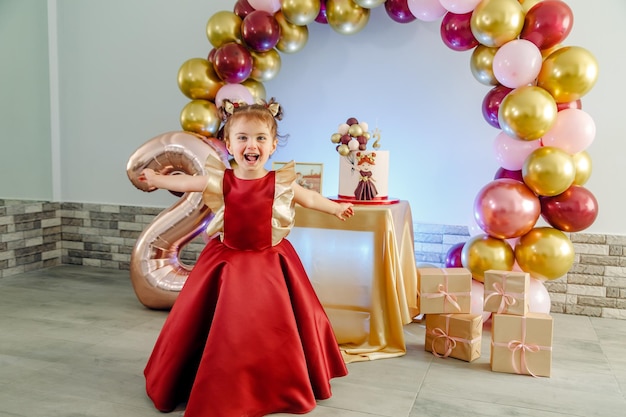 Divertida niña de dos años con ropa roja divirtiéndose frente a su pastel de cumpleaños. Hermosa sesión de fotos de cumpleaños de niña