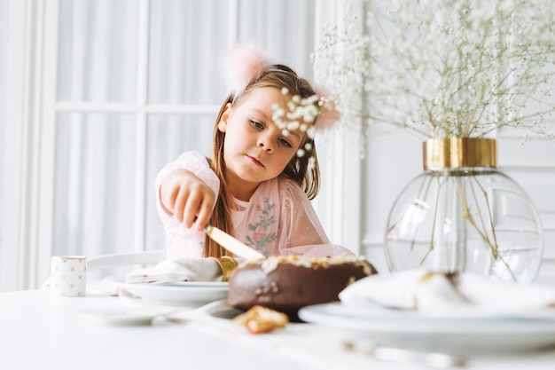 Divertida niña linda con el pelo largo en vestido rosa claro con pastel de chocolate en las manos en la mesa festiva en la luminosa sala de estar en la casa Navidad cumpleañera