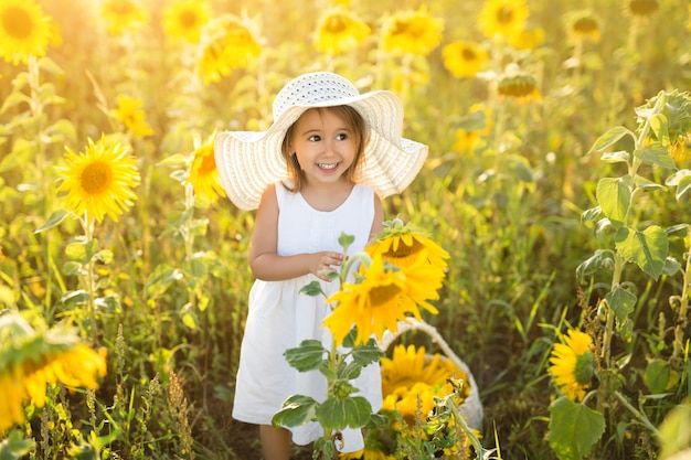 Una divertida niña linda con un sombrero en un campo de girasoles sostiene una flor en sus manos