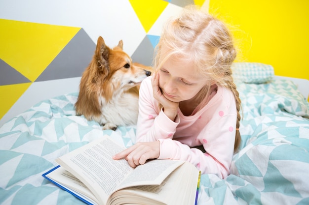 Divertida niña y perro corgi en la cama en la guardería leer un libro. educación en casa