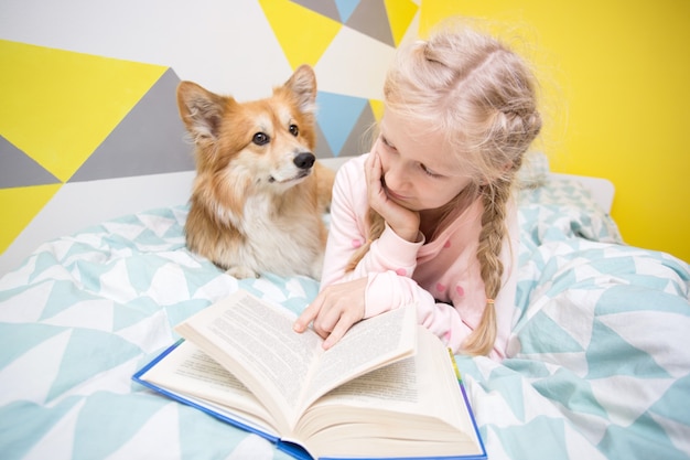 Divertida niña y perro corgi en la cama en la guardería leer un libro