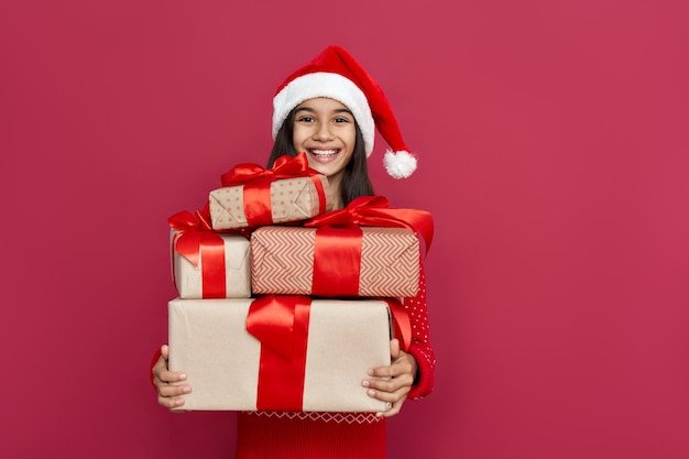 Divertida niña preadolescente latina india usa sombrero de Papá Noel mirando la cámara sosteniendo muchas cajas de regalos aisladas sobre fondo rojo Feliz Navidad presenta compras venta Feliz celebración de Año Nuevo 2021