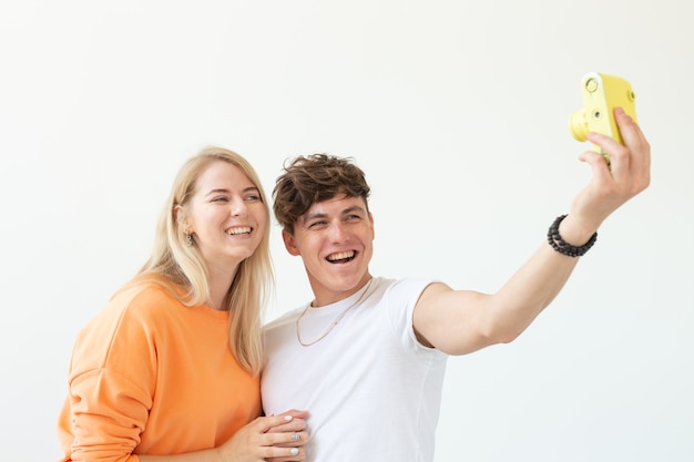 Divertida pareja de enamorados hombre lindo y mujer encantadora haciendo selfie en cámara de película amarilla vintage posando sobre una pared blanca. Amantes del concepto de fotografía y aficiones.