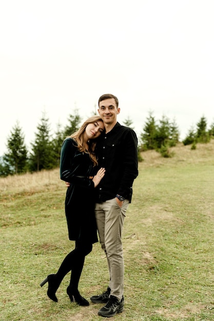 Divertida pareja joven en las montañas de otoño Un chico y una chica están parados cerca del arco de la boda bajo un gran árbol viejo contra el fondo de un bosque y picos montañosos al atardecer