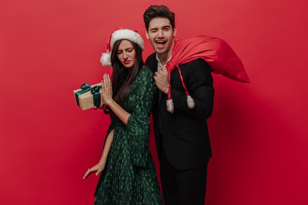 Divertida pareja de personas con trajes elegantes con atributos navideños posando emocionalmente aislado en la pared roja