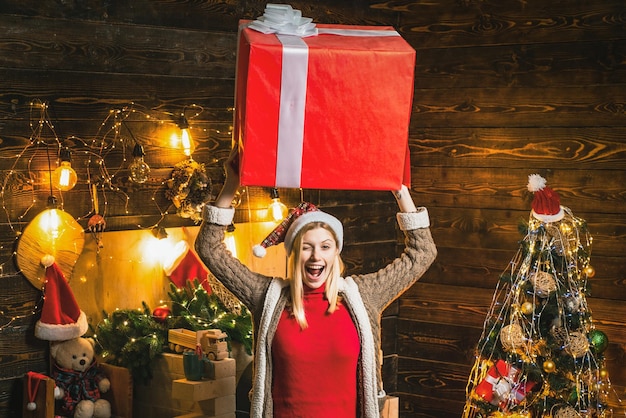 Divertida rubia con sombrero de Navidad y regalos celebrando las vacaciones de Navidad con caja grande en la cabeza.