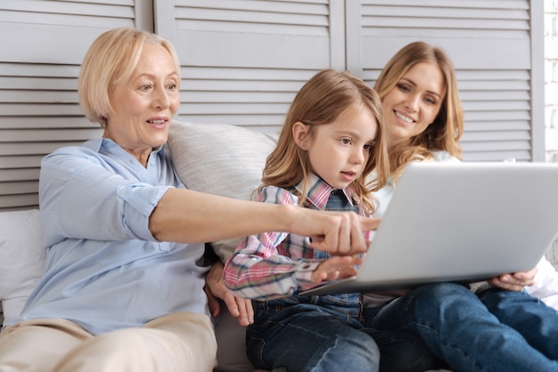 Divertida senhora idosa sentada ao lado do neto e da filha na cama e apontando para a tela do laptop enquanto a neta digita algo