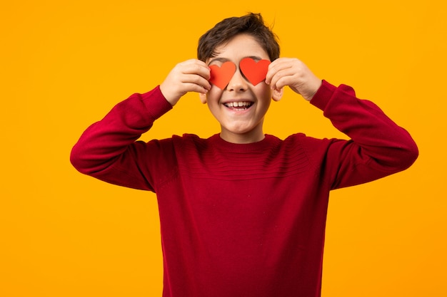 Divertido chico caucásico guapo con dos tarjetas de papel en forma de corazones para el Día de San Valentín en naranja