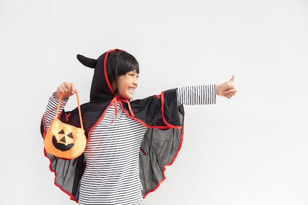 Divertido concepto de niño de halloween, niña linda con disfraz de fantasma de halloween asustadizo con fantasma de calabaza naranja en la mano, sobre fondo blanco