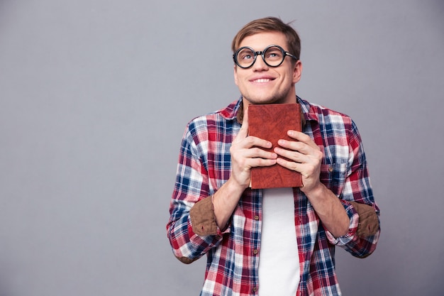 Divertido joven alegre en gafas redondas y camisa a cuadros sosteniendo libro sobre pared gris