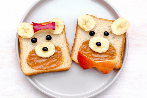 Divertido lindo beardog cara sándwich tostadas pan con mantequilla de maní plátano y manzana Niños niños bebé dulce postre saludable desayuno almuerzo comida arte en platewhite fondo cerrar vista superior