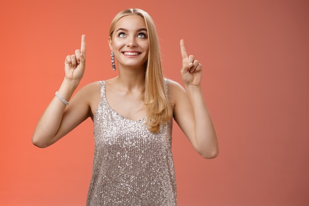 Divertido lindo concurso elegante mulher europeia loira longo penteado em prata brilhante vestido de noite olhar apontando para cima olhar curioso excitação ver a coisa desejada, em pé fundo vermelho alegre.