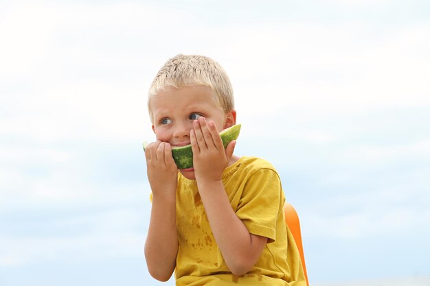 Divertido niño rubio bronceado en la costa, sosteniendo una jugosa rebanada de sandía y comiéndola con gusto. Concepto de verano.