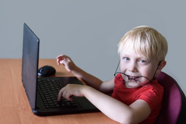 Divertido niño rubio con gafas se sienta en una computadora portátil, Internet y preescolar