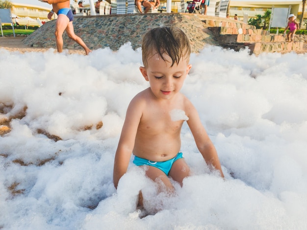 Divertido retrato de niño pequeño cubierto de espuma de jabón jugando en la playa durante la fiesta disco