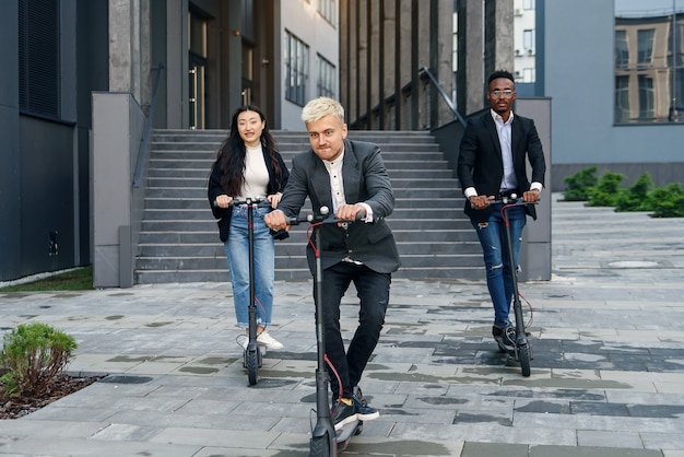 Divertidos colegas de oficina multirraciales alegres que tienen competencia de carreras en scooters eléctricos cerca de moderno