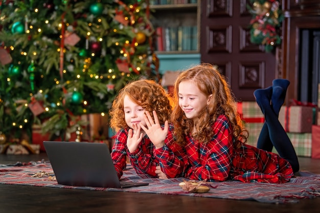 Divertidos niños pelirrojos junto al árbol de Navidad y los regalos se comunican online a través de un portátil.