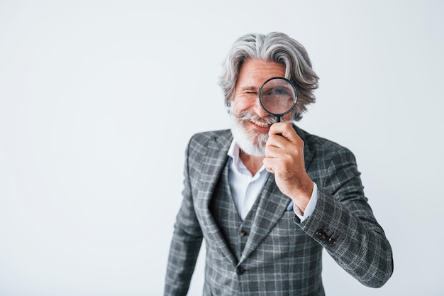 Divertindo-se com lupa Homem moderno elegante sênior com cabelos grisalhos e barba dentro de casa