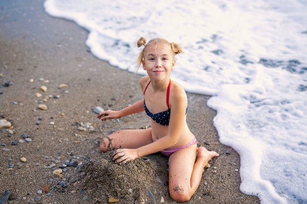 Divirtiéndose y alegría. Chica encantadora rubia con dos trenzas jugando con arena de mar.