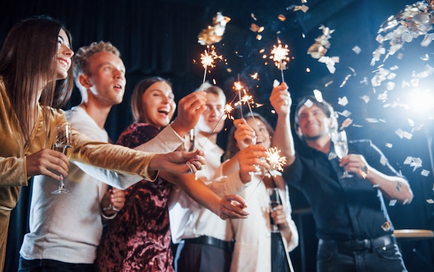 Divirtiéndose con bengalas. El confeti está en el aire. Grupo de amigos alegres celebrando el año nuevo en el interior con bebidas en las manos.