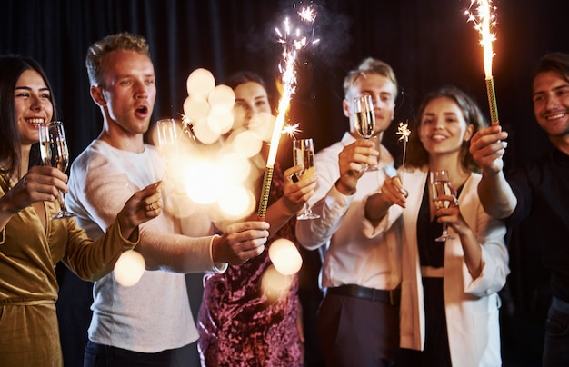 Divirtiéndose con bengalas. Grupo de amigos alegres celebrando el año nuevo en el interior con bebidas en las manos.