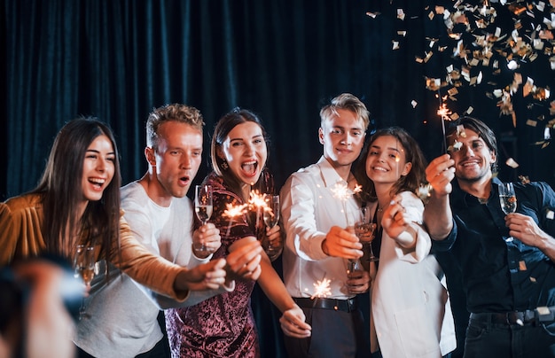 Divirtiéndose con bengalas. Grupo de amigos alegres celebrando el año nuevo en el interior con bebidas en las manos.