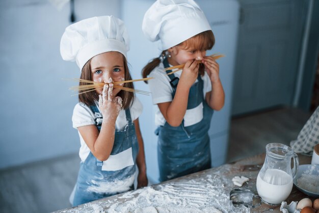 Divirtiéndose con espaguetis. Niños de la familia en uniforme de chef blanco preparando la comida en la cocina.